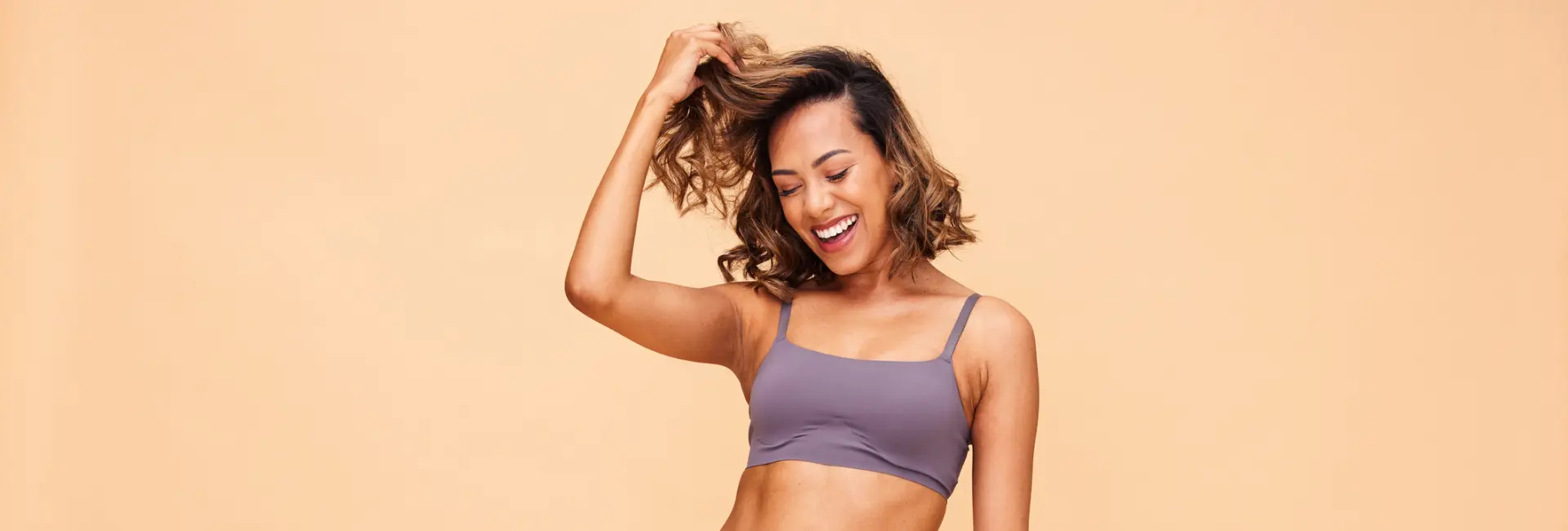 A woman in a gray sports bra smiling with her eyes closed, holding her hair up against a beige background.