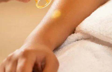 A hand applies a drop of oil onto a person's arm, which rests on a towel-covered surface.