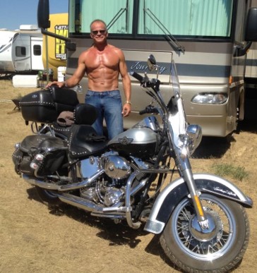 A man stands shirtless beside a parked motorcycle, with an RV in the background on a grassy area.