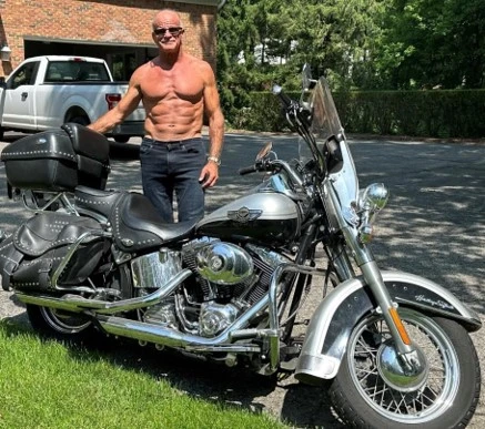 A shirtless man stands next to a parked motorcycle in a driveway with a truck and greenery in the background.