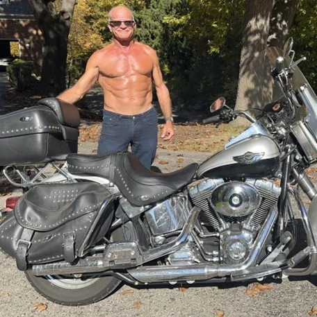 A shirtless man with sunglasses stands behind a parked motorcycle on a sunny day.