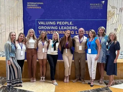 A group of eleven people stand in front of a blue banner at the International Maxwell Conference, posing for a group photo.