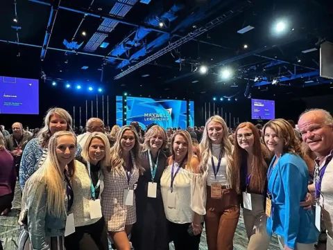 A group of people pose for a photo at a conference event, standing indoors with a stage and "Maxwell Leadership" displayed in the background.
