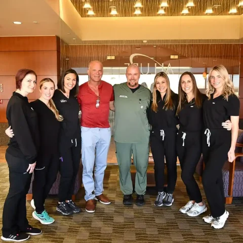 A group of eight adults, some in scrubs and others in casual attire, stand together and smile in a modern lobby area.