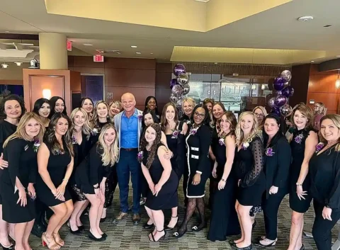 A group of women and one man pose together indoors, all dressed in black attire, with purple and silver balloons in the background.