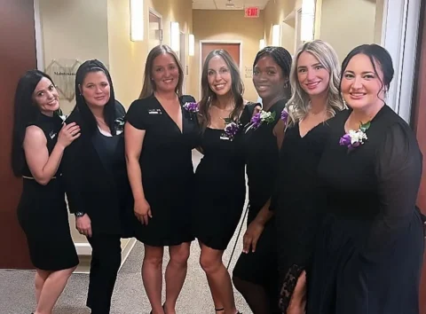 Seven women dressed in black outfits with name tags and purple and white corsages stand in a hallway, posing for a group photo.