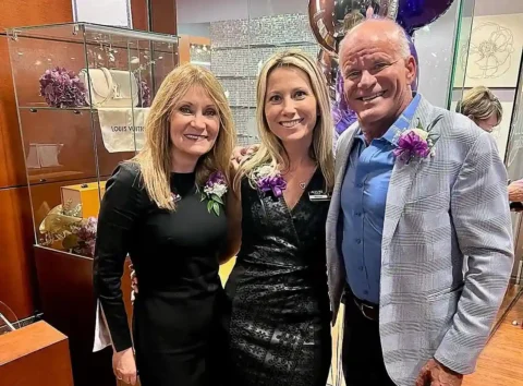 Three adults, two women and one man, dressed formally and wearing corsages, pose together and smile in a retail store decorated with flowers and balloons.