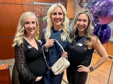 Three women smiling indoors; one is visibly pregnant, and they all wear name badges. Purple balloons are visible in the background.
