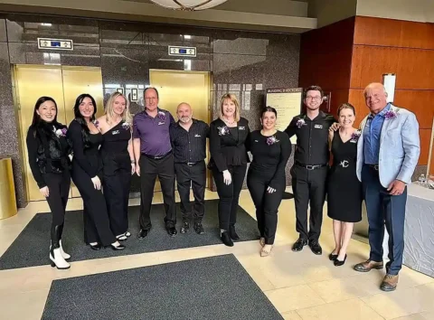 A group of nine people dressed in formal attire pose together and smile for a photo in a building lobby near elevators.