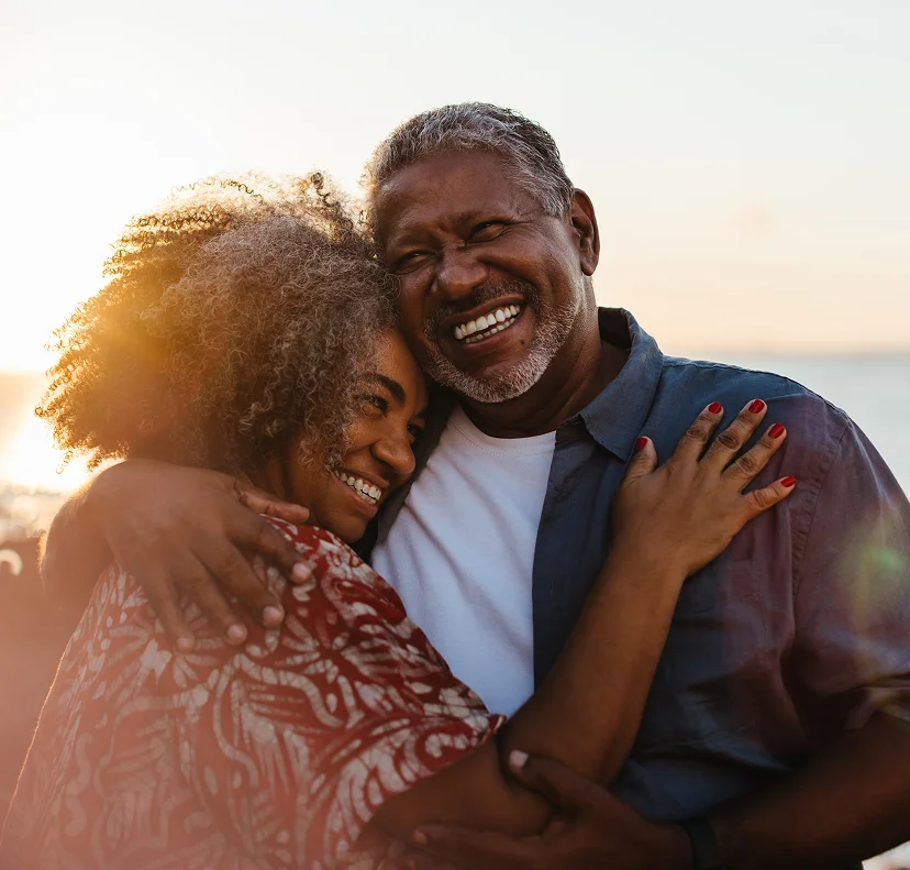 A smiling couple embraces outdoors with sunlight in the background. The woman wears a patterned shirt, and the man wears a blue shirt.