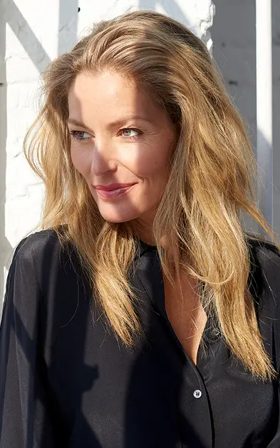 Person with long blonde hair and a black shirt smiling slightly, looking to the side, with sunlight casting shadows in the background.