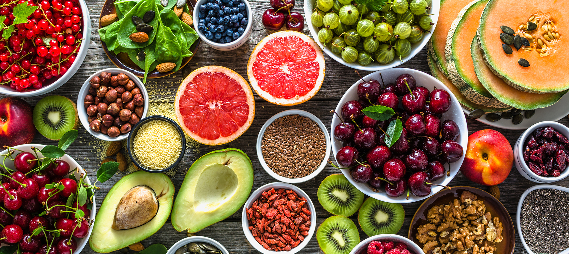 Assorted fruits, nuts, and seeds arranged in bowls on a wooden surface, including cherries, avocado, kiwis, blueberries, and grapefruit.