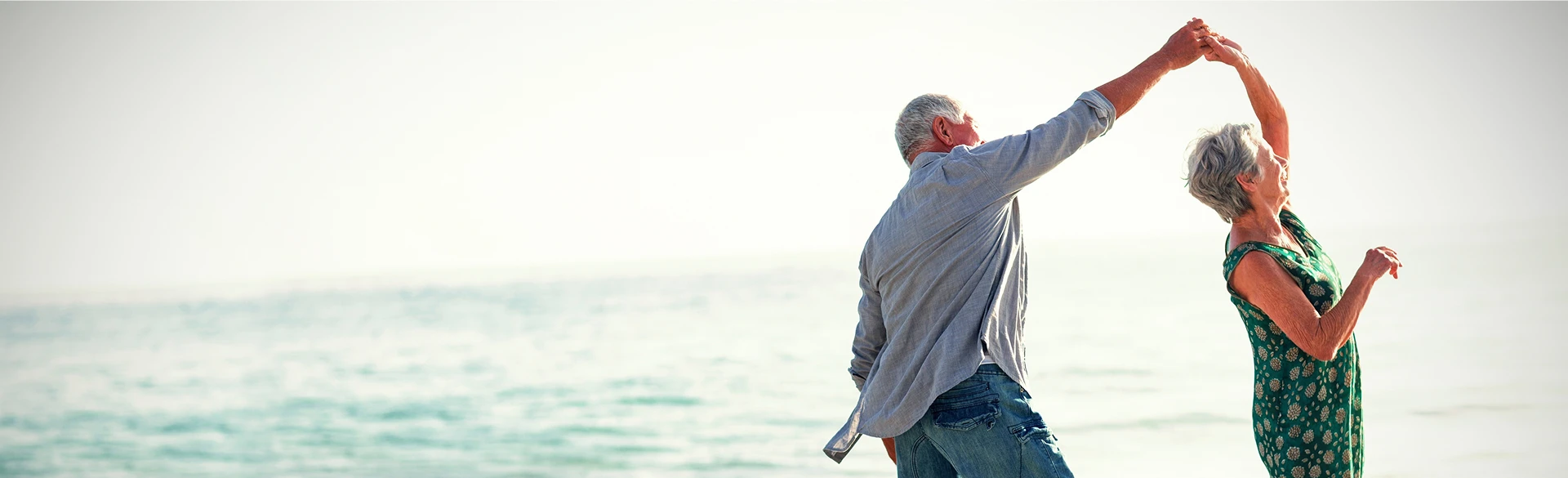 An older couple dances together on a beach near the ocean, with the man twirling the woman under his arm.