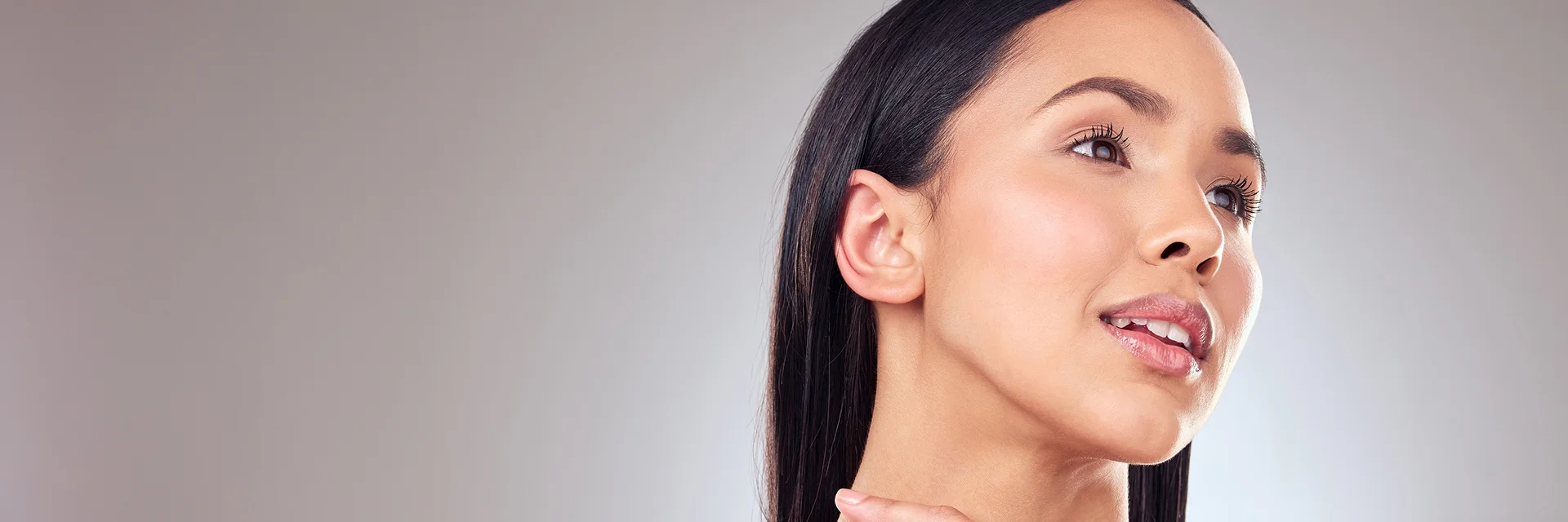 A woman with clear skin and straight dark hair looks upward, gently touching her neck, against a plain gray background.