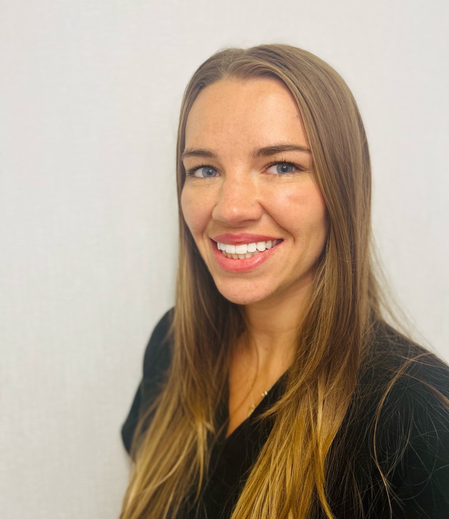 A woman with long, straight, light brown hair and a black top smiles at the camera against a plain, light-colored background.