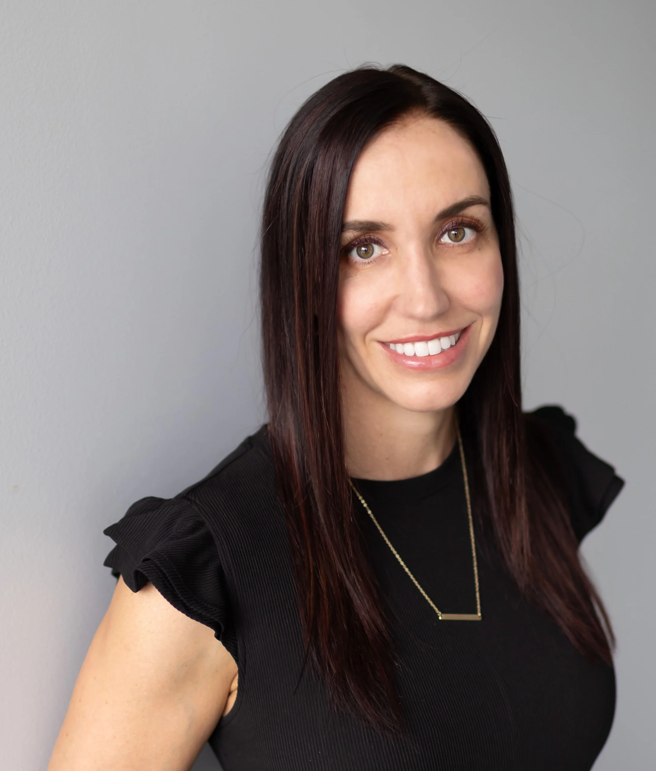 A woman with straight dark hair and a black dress stands against a gray background, smiling at the camera.