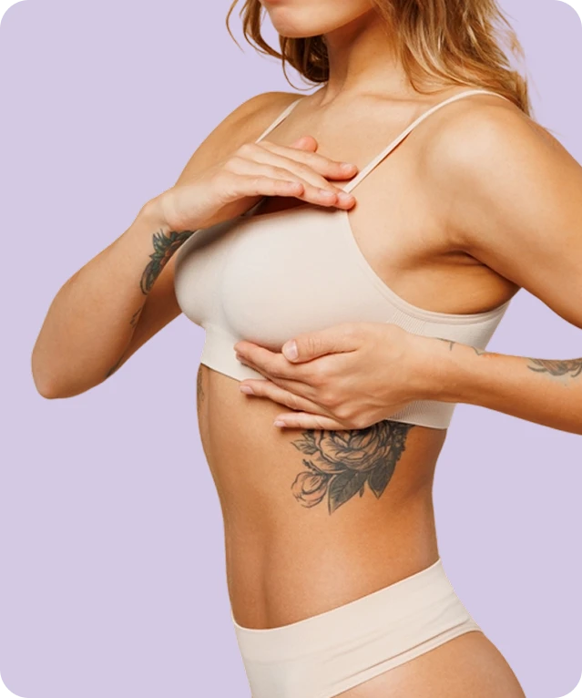 Woman in beige underwear performing a breast self-exam, with visible tattoos on her arm and torso, against a light purple background.