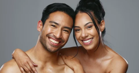 A man and woman stand close together, smiling at the camera with arms around each other, against a plain grey background.