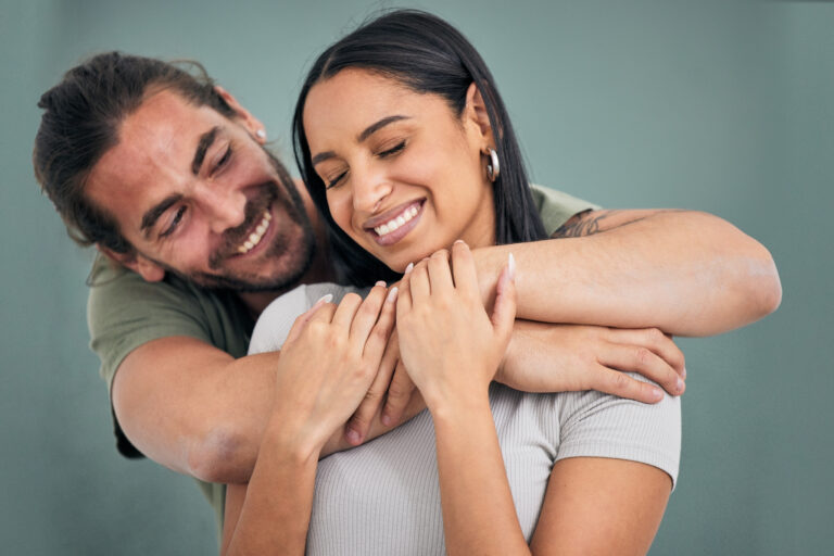 A man hugs a smiling woman from behind against a plain background. Both appear relaxed and happy.