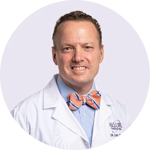 A man wearing a white medical coat and a colorful bow tie, smiling at the camera against a plain background.