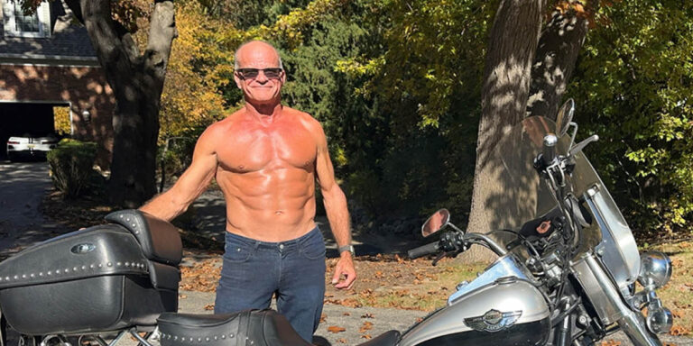 A shirtless older man with sunglasses stands outdoors next to a parked motorcycle on a sunny day with trees and a house in the background.