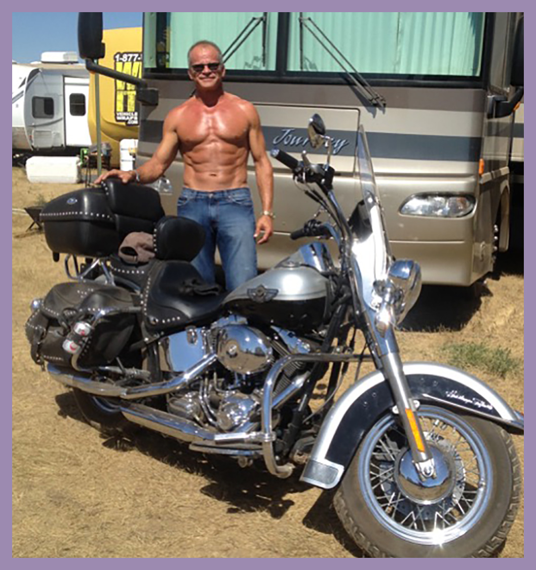 A shirtless man wearing sunglasses stands next to a parked motorcycle in front of a large RV on a sunny day.