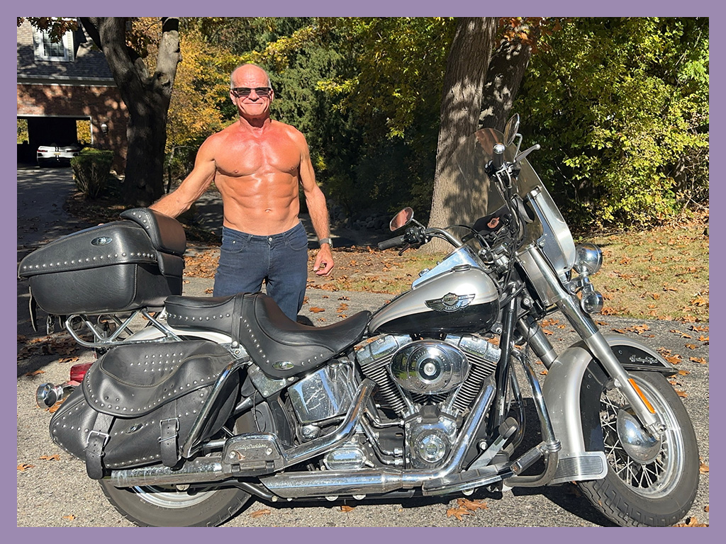 A shirtless man wearing sunglasses stands outdoors next to a large black and silver motorcycle on a sunny day, with trees and a house in the background.
