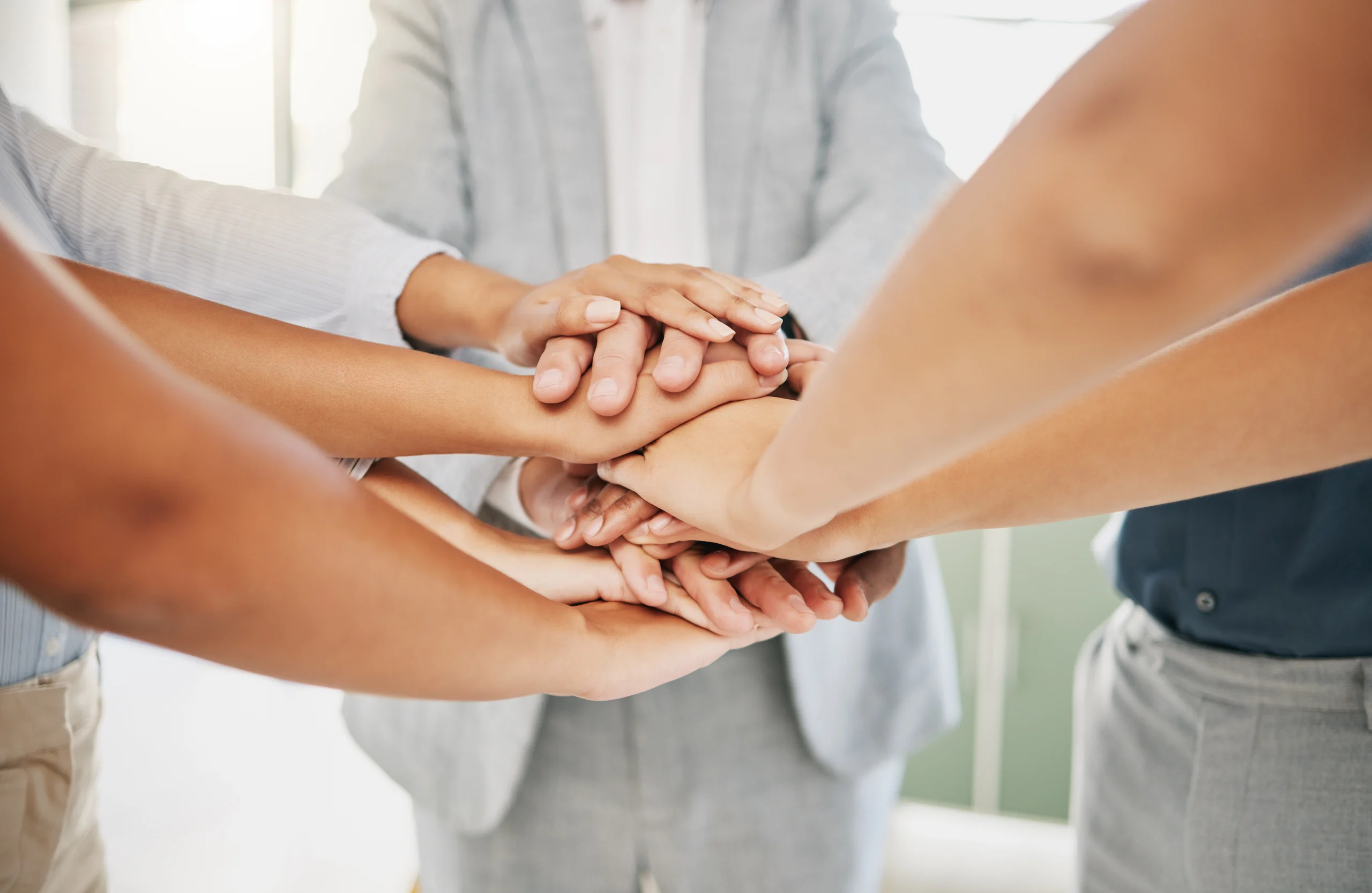A group of people standing in a circle stack their hands together in the center, symbolizing teamwork and unity.