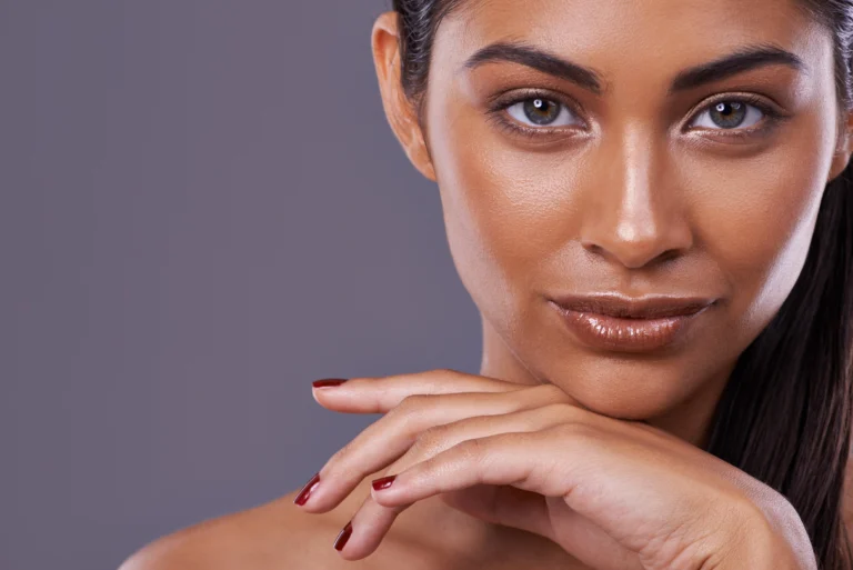 A woman with brown skin and straight dark hair poses with her chin resting on her hand, displaying neutral makeup and maroon nail polish against a gray background.