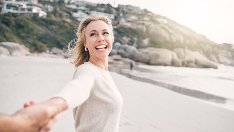 A woman smiling and holding someone’s hand while walking on a sandy beach with rocky hills and houses in the background.