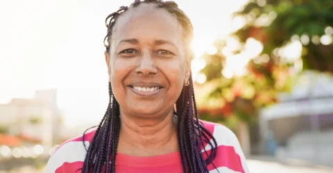 A woman with braided hair and a pink striped shirt smiles outdoors with sunlight in the background.