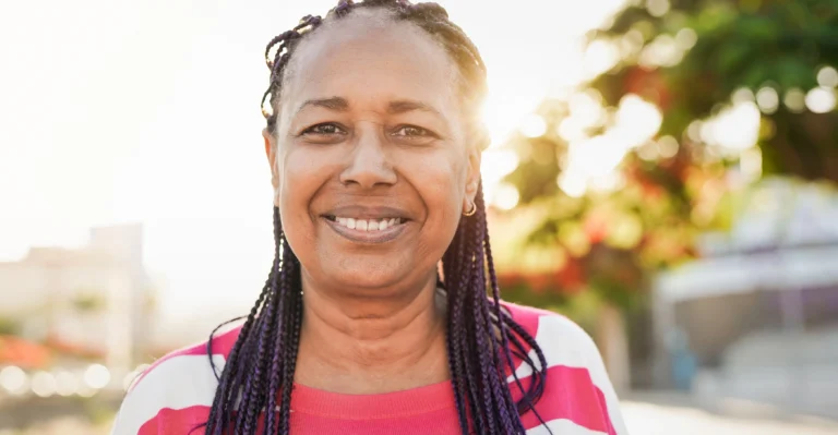A woman with braided hair and a pink striped shirt smiles outdoors with sunlight in the background.