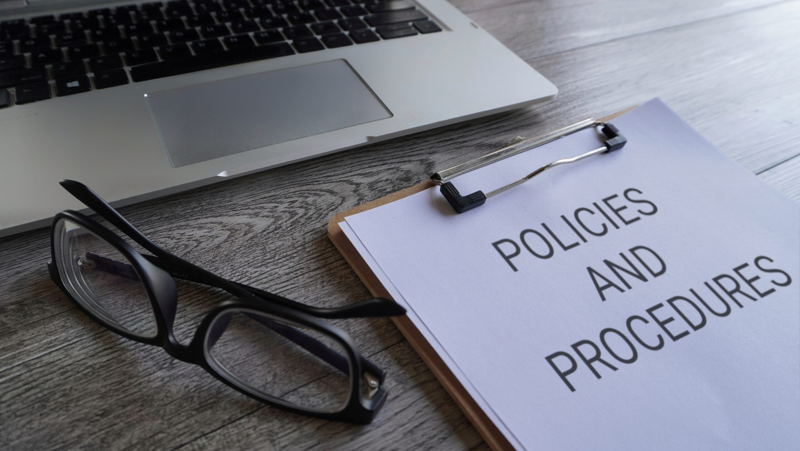 A pair of eyeglasses, a laptop, and a clipboard holding a document titled "POLICIES AND PROCEDURES" on a wooden desk.