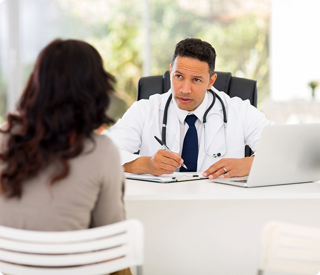 A doctor wearing a white coat and stethoscope sits at a desk, taking notes while speaking with a patient across from him.