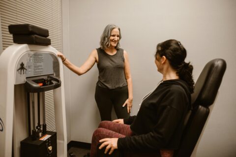 Two women are in a gym room; one is standing and smiling, while the other sits on an exercise machine, both are wearing workout clothes.