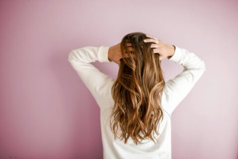 Person with long, wavy, brown hair wearing a white top stands facing a light pink wall, with both hands placed on the back of their head.