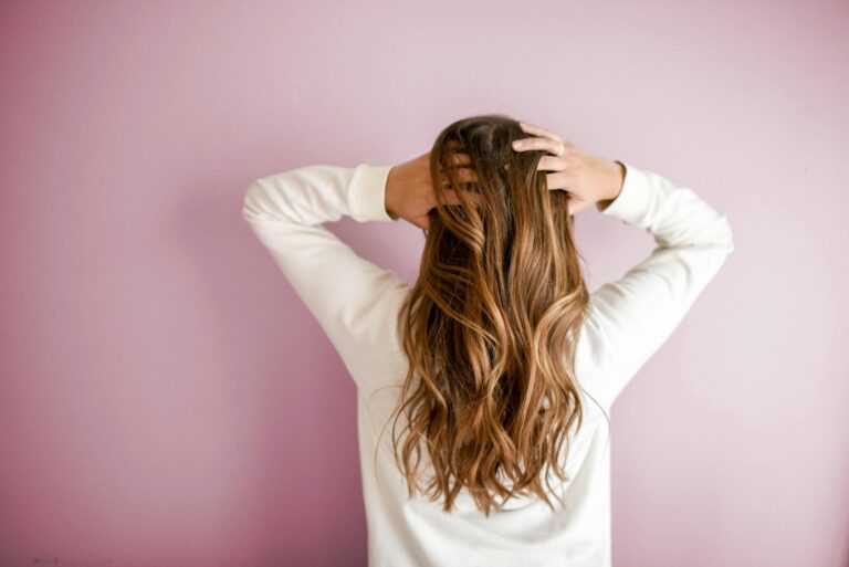 Person with long, wavy, brown hair wearing a white top stands facing a light pink wall, with both hands placed on the back of their head.