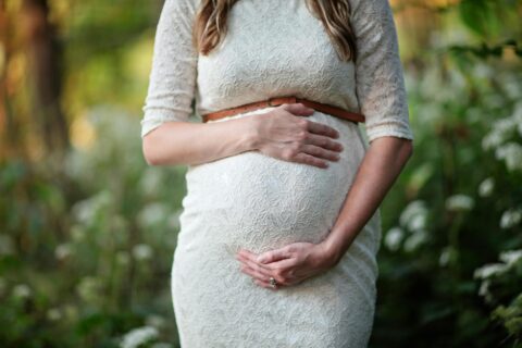 A pregnant woman in a white lace dress stands outdoors, holding her belly with both hands.