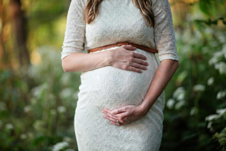 A pregnant woman in a white lace dress stands outdoors, holding her belly with both hands.
