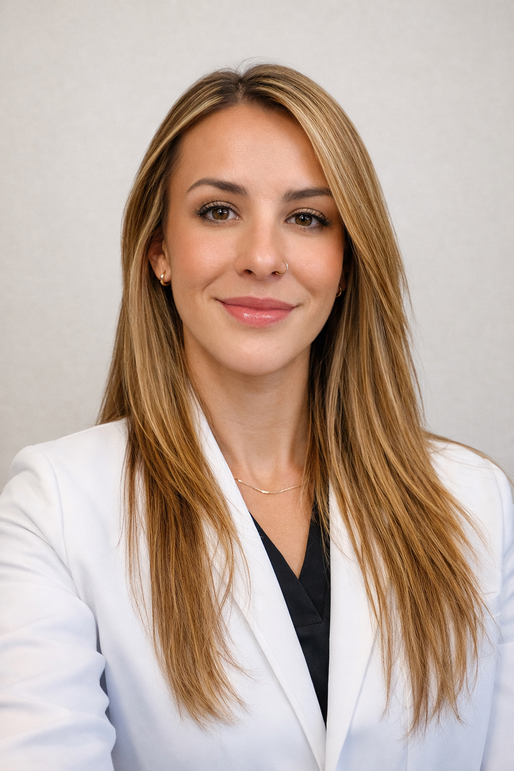 A woman with long straight blonde hair wearing a white lab coat and black top, smiling in front of a plain light background.