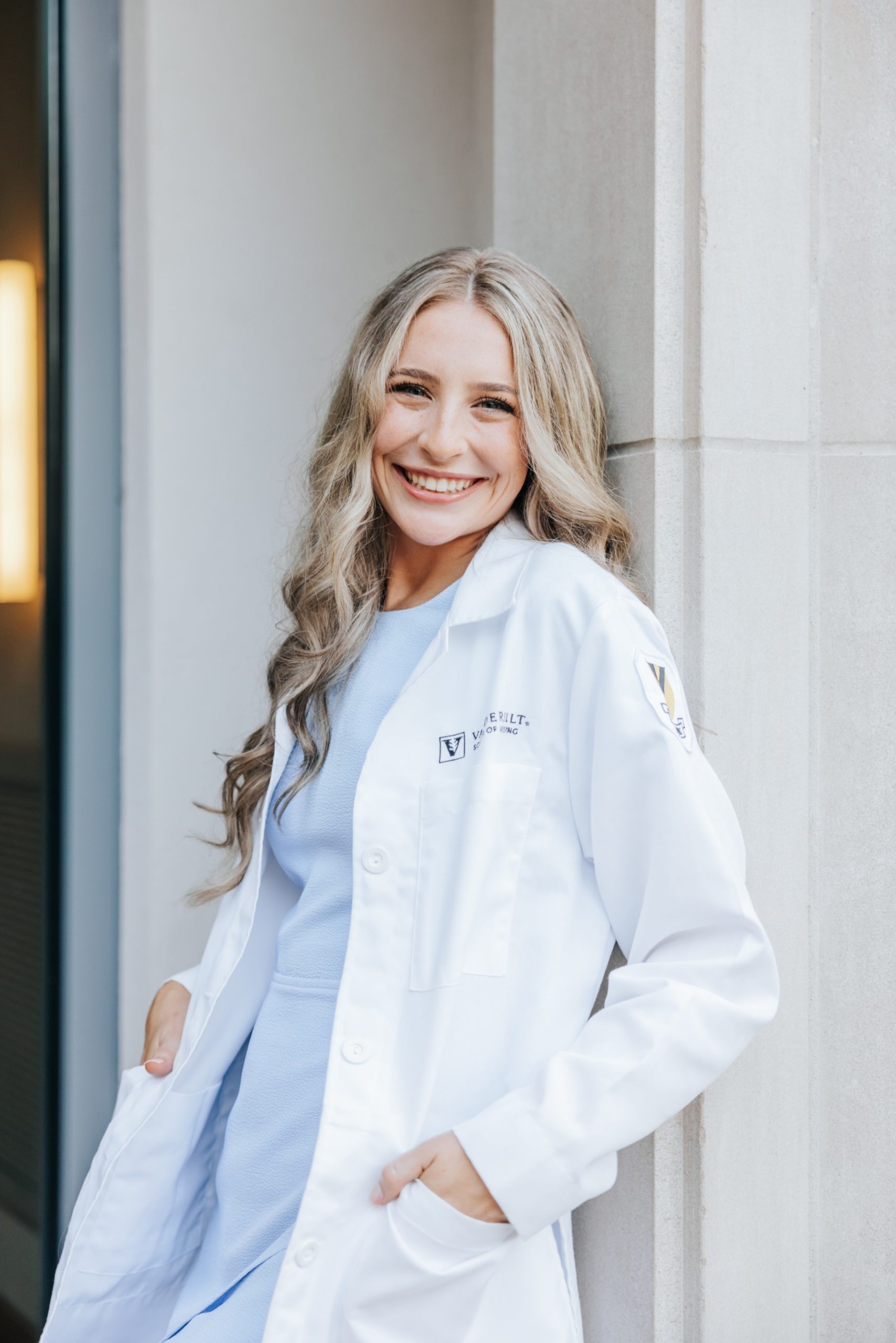 A woman with long wavy hair, wearing a white lab coat and light blue clothing, stands smiling with her hands in her pockets against a light-colored wall.